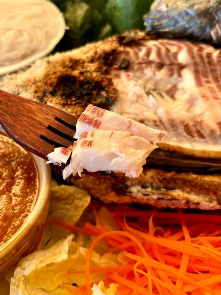 A wooden fork holds a juicy piece of grilled fish flesh besides a bowl of seafood dipping sauce.