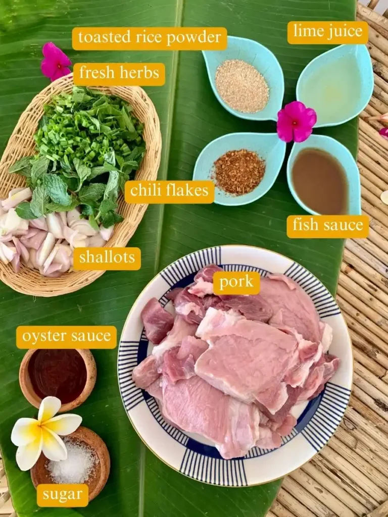 Top-view of ingredients for nam tok moo salad laid out on a banana leaf including sliced pork, toasted rice powder, herbs, shallots, chili flakes, lime juice, fish sauce, oyster sauce, and sugar, ready for Thai cooking.