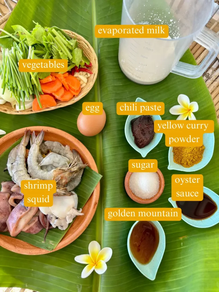 Top-view of ingredients for pad pong karee seafood laid out on banana leaves, including shrimp, squid, vegetables, evaporated milk, egg, yellow curry powder, chili paste, sugar, oyster sauce, and golden mountain.