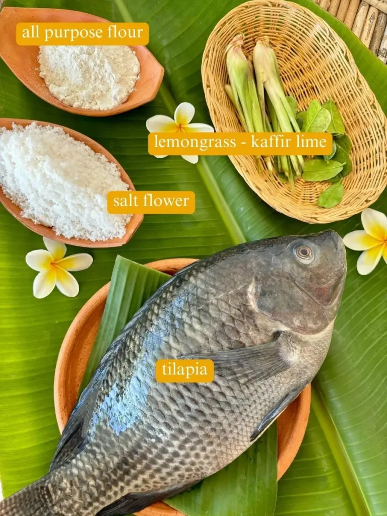 Fresh tilapia presented on a banana leaf with lemongrass and kaffir lime leaves, with bowls of all-purpose flour and salt flour.