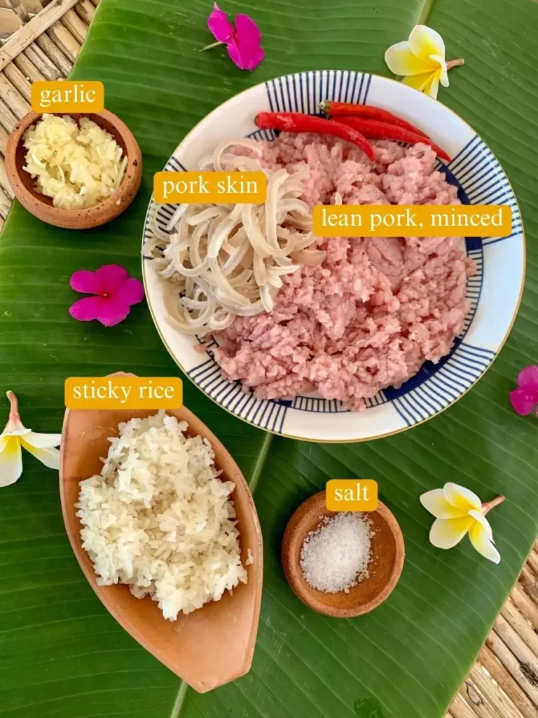 Ingredients for som moo displayed on a banana leaf: minced lean pork, sliced pork skin, crushed garlic, sticky rice, and salt.