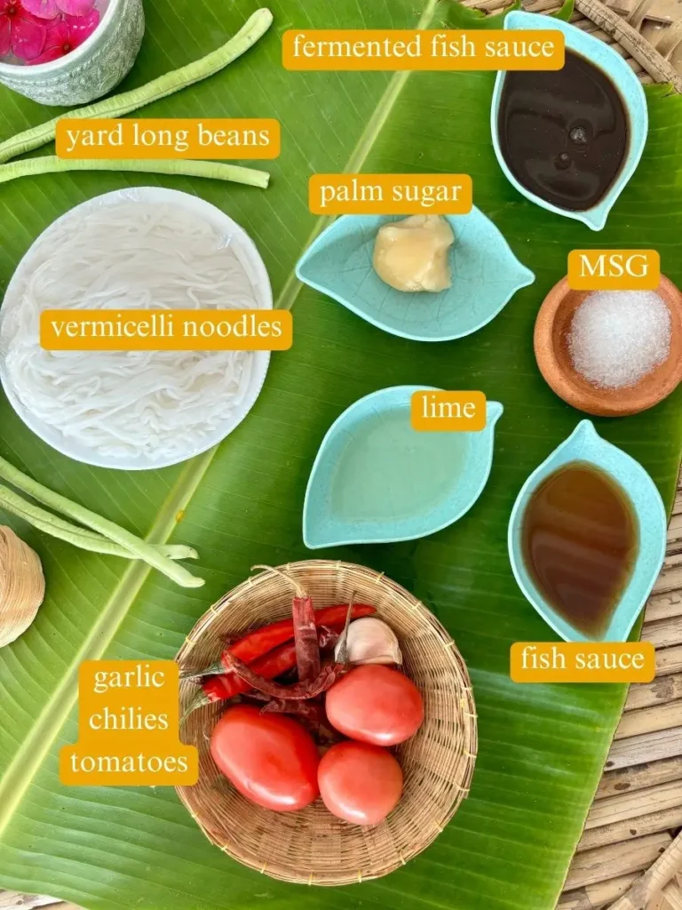 Top-view of ingredients for thum khao poon laid out: vermicelli noodles, yard long beans, palm sugar, lime, MSG, fermented fish sauce, garlic, chilies, and tomatoes, ready for preparation.