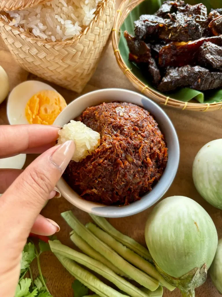 Hand scooping sticky rice with spicy jeow bong, a Lao chili paste, accompanied by fresh vegetables and beef jerky.