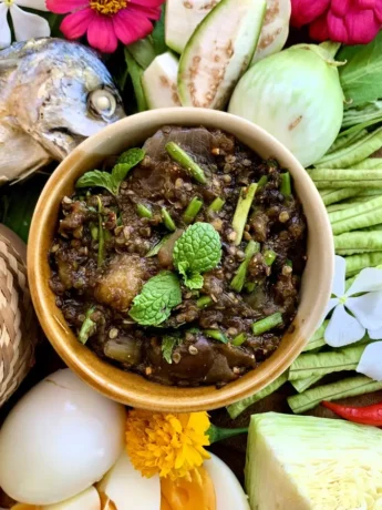 Lao jeow mak keua served in a bowl with sticky rice, fresh vegetables, hard-boiled eggs, and grilled fish, garnished with mint leaves.