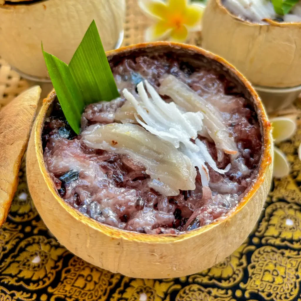 Khao lam, Thai sticky rice in bamboo dessert, with black beans topped with young coconut meat presented in a coconut shell.