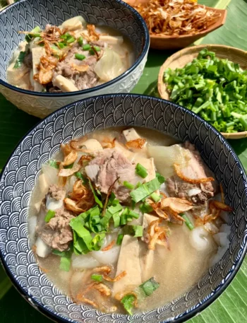 Steaming bowl of khao piak sen, a Lao noodle soup with pork, homemade noodles, garnished with fried shallots and green onions on a banana leaf background.