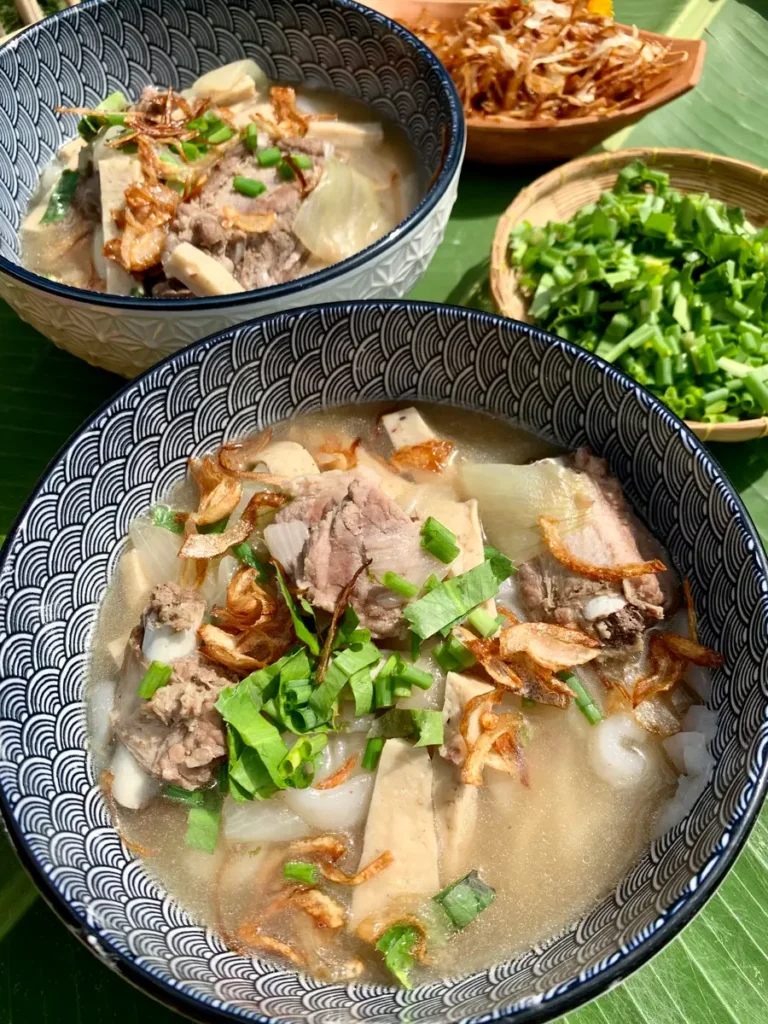 Steaming bowl of khao piak sen, a Lao noodle soup with pork, homemade noodles, garnished with fried shallots and green onions on a banana leaf background.