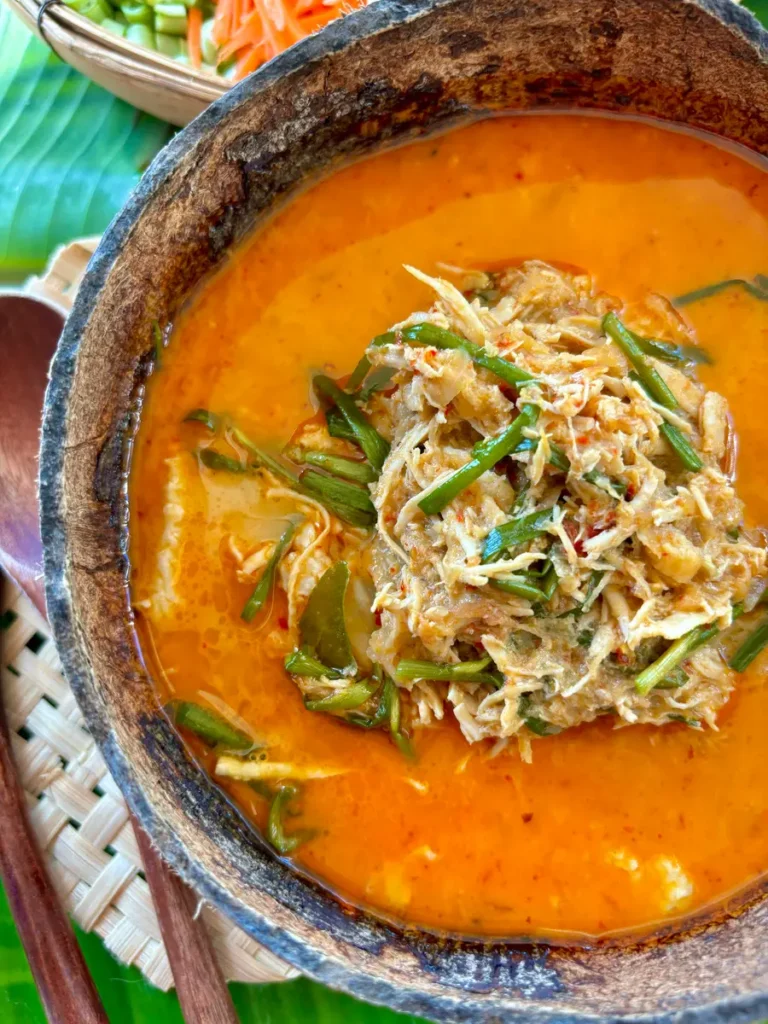 Authentic khao poon, Lao coconut noodle soup, with a red curry broth, garnished with green onions and shredded chicken, served in a coconut shell.