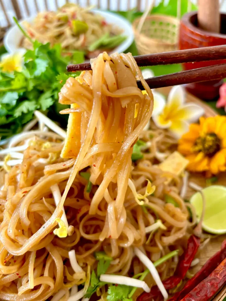 Close-up of kua mee noodles lifted on chopsticks, highlighting the rice noodles coated in sauce, garnished with fresh herbs.