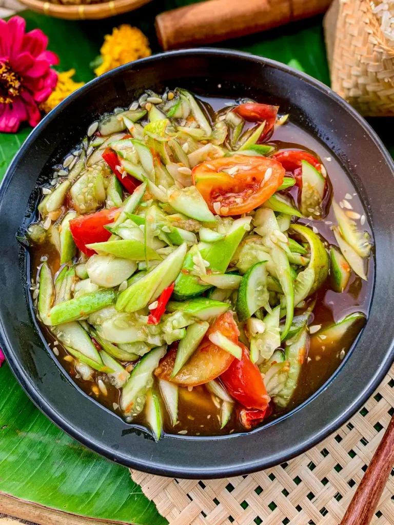 Lao cucumber salad (thum mak thang) with green mango, tomato, and chili in a black bowl over banana leaf.