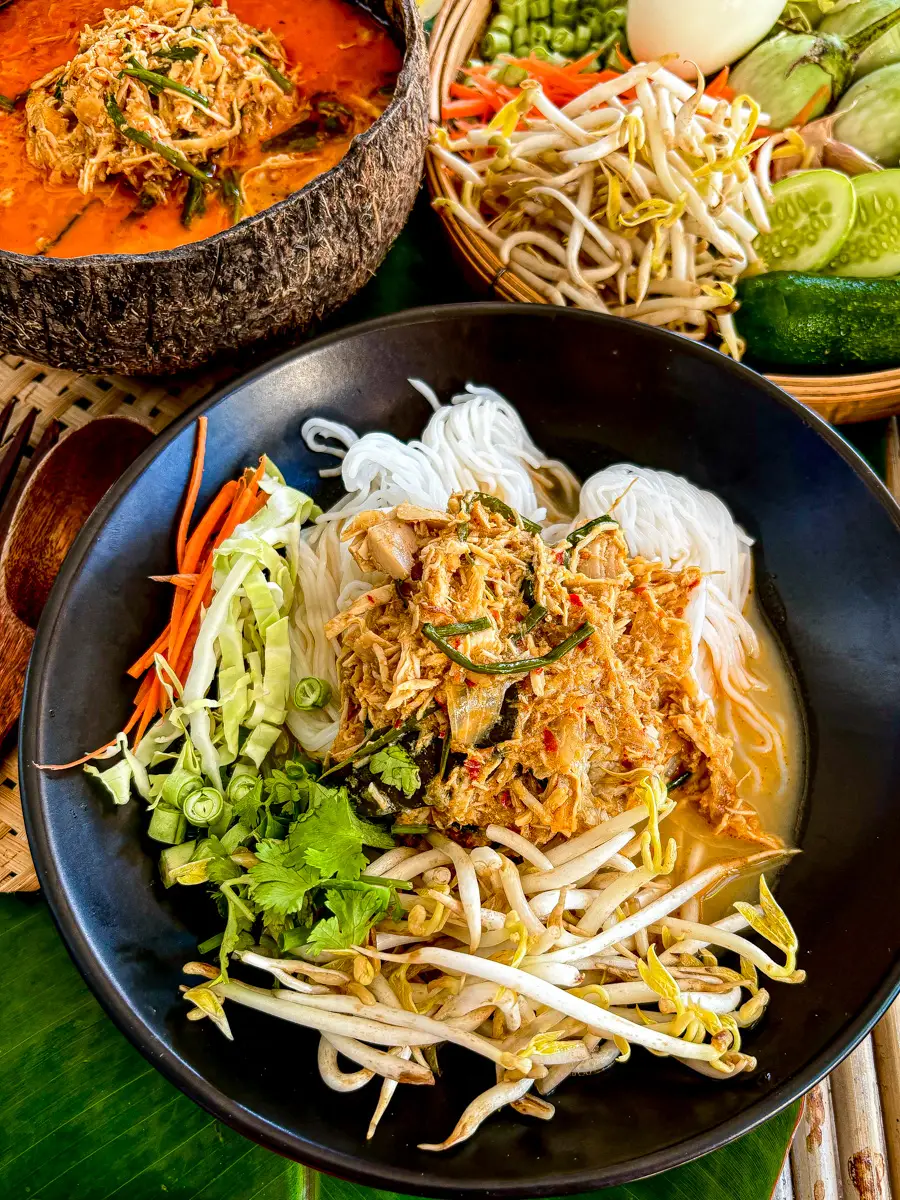 Lao chicken curry noodle soup (khao poon) with rice noodles, herbs, and raw vegetables arranged on the side.