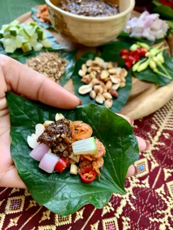 Handful of miang kham on a wild pepper leaf with assorted Thai garnishes like roasted peanuts, dried shrimp, diced lime, and chili, set on a traditional Thai fabric.