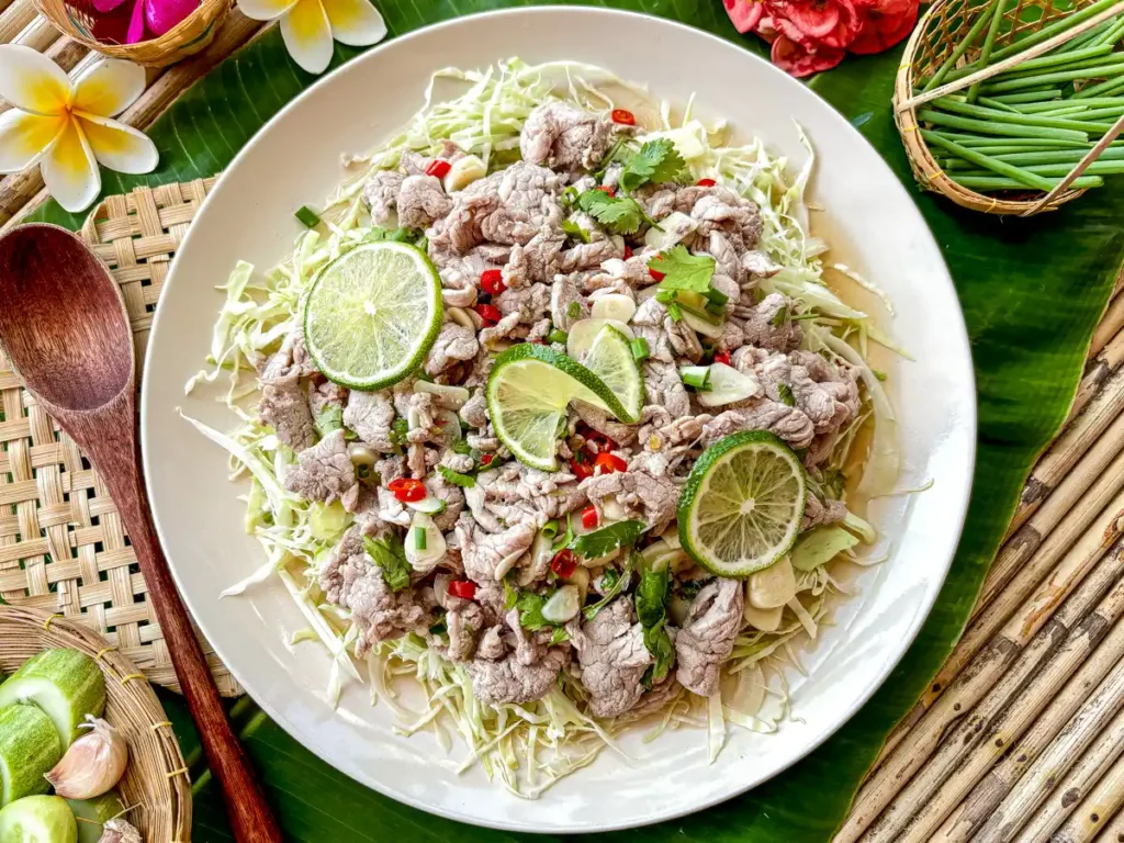 Moo manao, Thai lime pork salad topped with garlic, chilies, and herbs on shredded cabbage, surrounded by tropical flowers and utensils.
