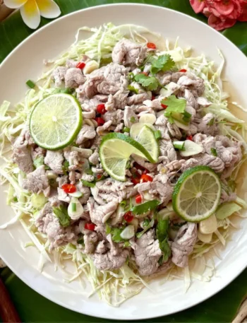 Authentic moo manao Thai salad with pork, shredded cabbage, fresh lime, and spicy chili garnish in a white dish.