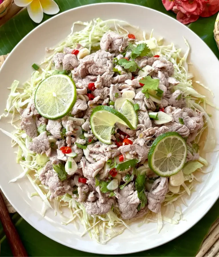 Authentic moo manao Thai salad with pork, shredded cabbage, fresh lime, and spicy chili garnish in a white dish.