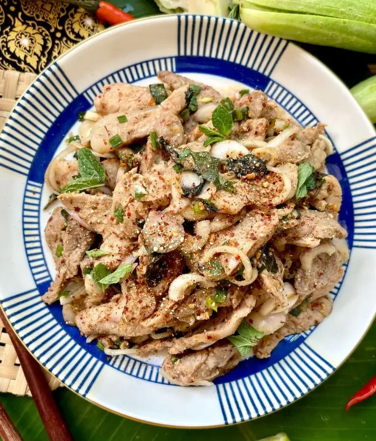 Authentic Thai nam tok moo recipe, a spicy grilled pork salad with mint, shallots, and chili flakes, served in a blue and white striped plate for a Thai meal.