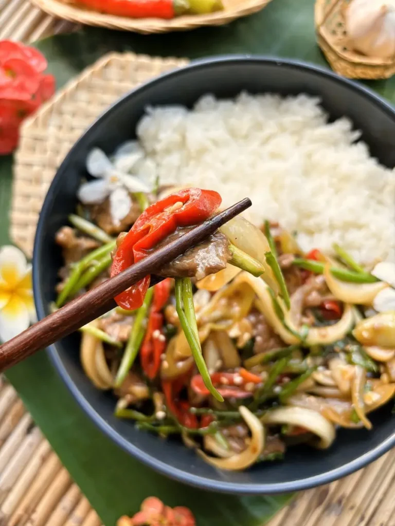 Close-up of Thai chili beef with crisp chilies and spring onions, picked up with chopsticks over steamed rice.