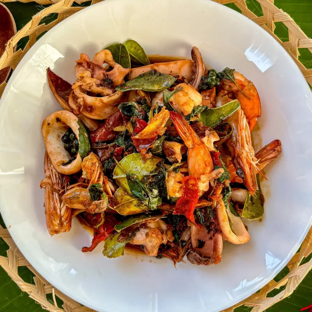 Pad cha talay, Thai stir-fried seafood with prawns, squid, red chili, green peppercorn, and basil on a white dish.