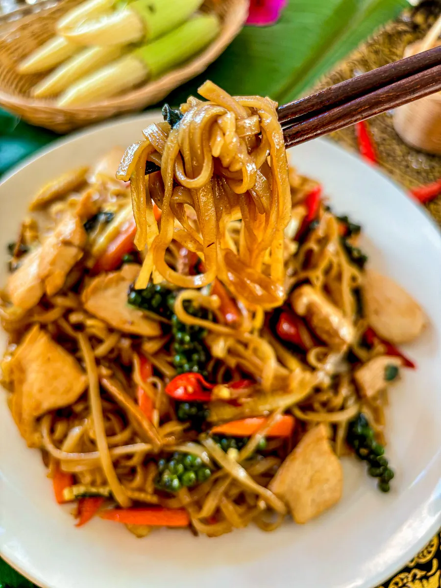 Pad kee mao noodles with chicken, baby corn, and chilies, topped with green peppercorns, captured mid-bite with chopsticks.