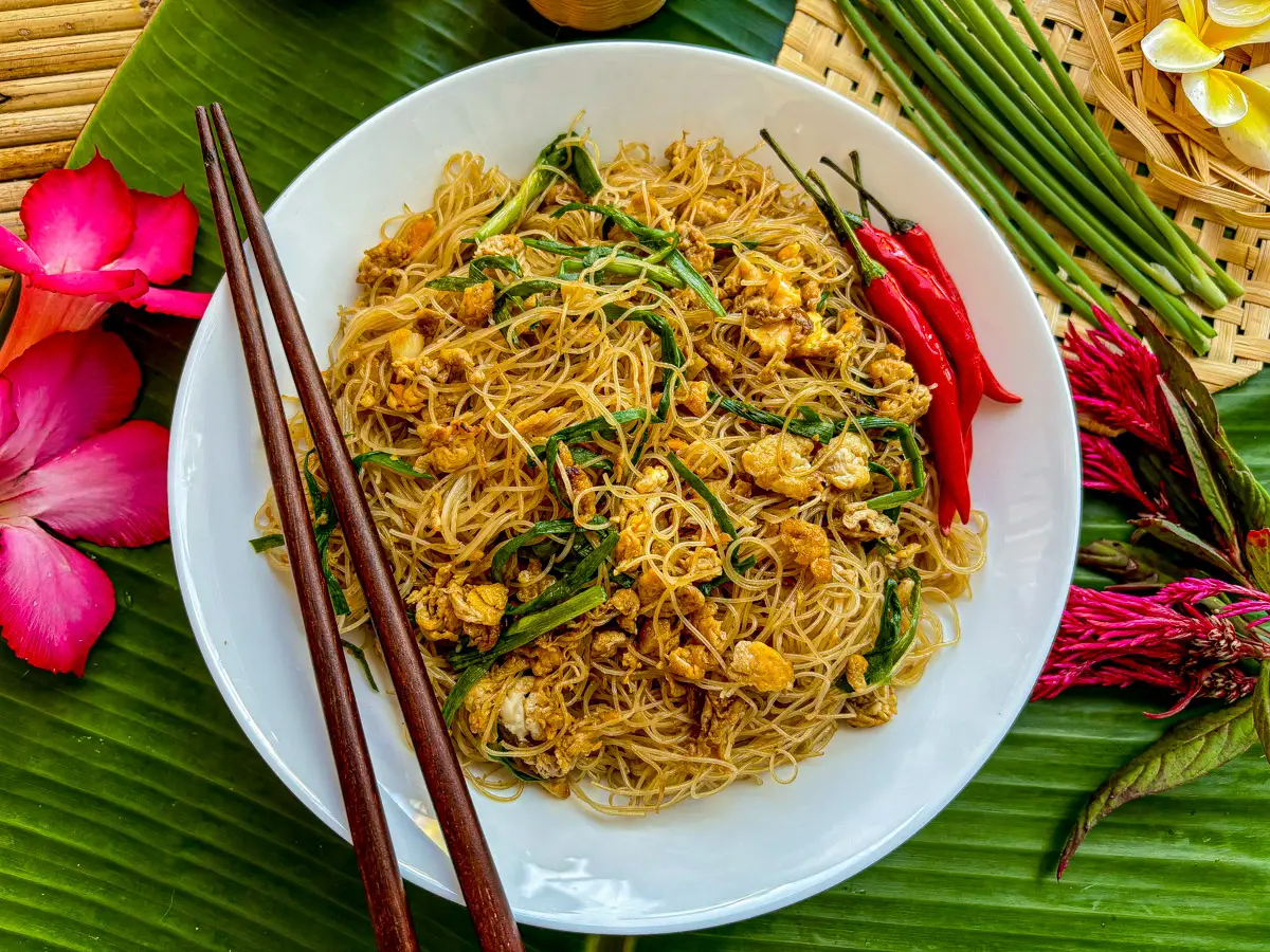 Pad mee, Thai stir-fried sen mee noodles on a white plate with chilies and garlic chives, styled with tropical flowers.