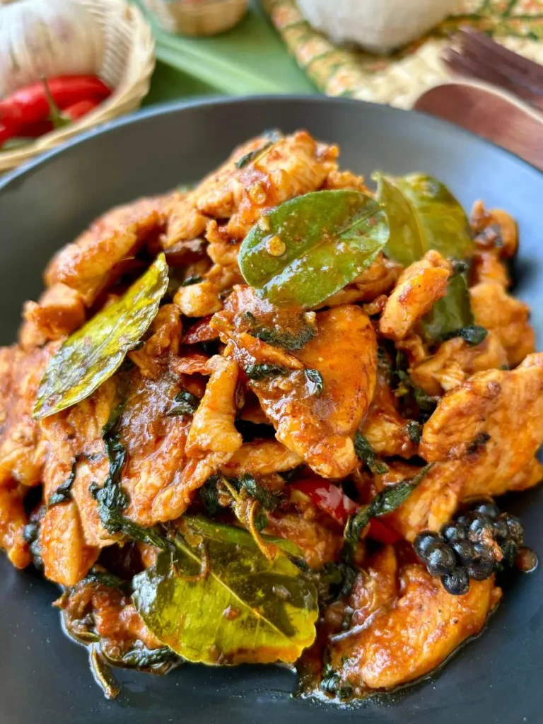 Close-up of pad ped chicken, a spicy red curry paste stir-fry featuring tender chicken pieces with black peppercorns, chilies, and fresh herbs and spices.