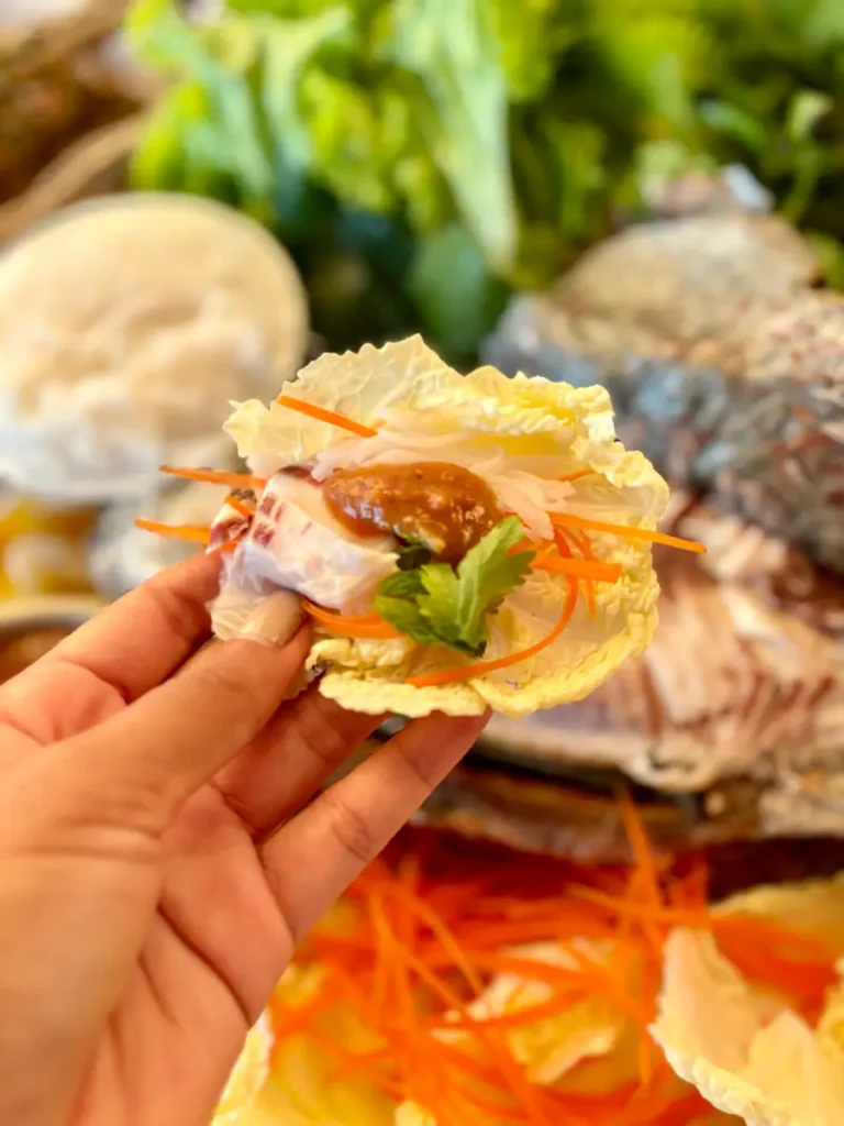 A hand presenting a lettuce wrap filled with grilled fish, vegetables, and rice vermicelli.