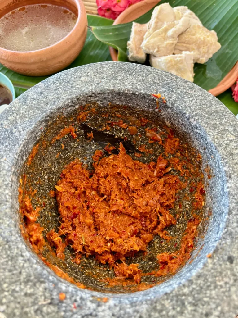 Red curry paste for khao poon in a stone mortar. In the distance is a bowl of chicken broth and boiled chicken.