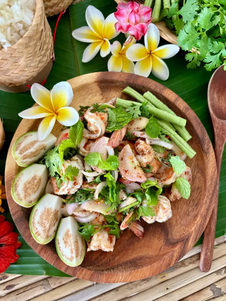 Shrimp larb salad presented in a wooden bowl on a banana leaf, accompanied by sticky rice and fresh herbs.