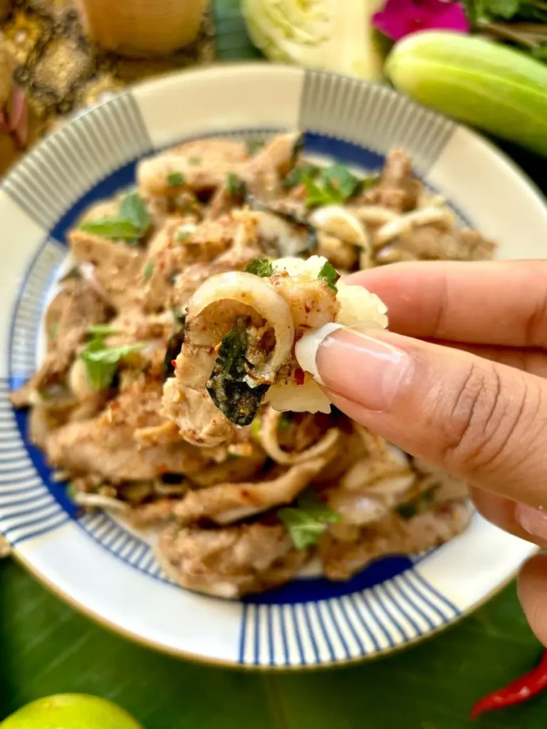 Close-up of a hand-picking up a piece of moo nam tok, showing the tender grilled pork with a sprinkle of fresh herbs.