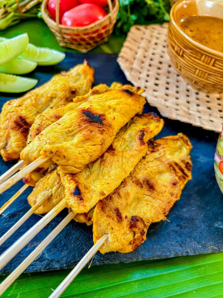 Thai satay peanut sauce served in a rustic brown bowl with grilled chicken skewers and cucumber salad on the side.