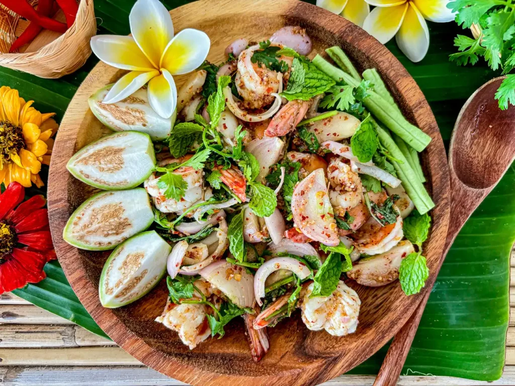 Thai shrimp larb in a wooden bowl with fresh herbs and Thai dressing on a banana leaf.