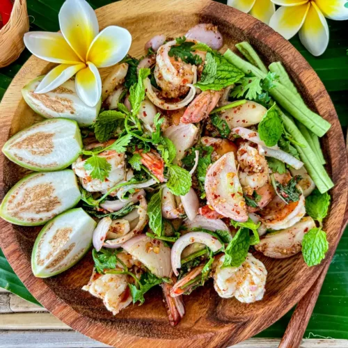 Thai shrimp larb in a wooden bowl with fresh herbs and Thai dressing on a banana leaf.