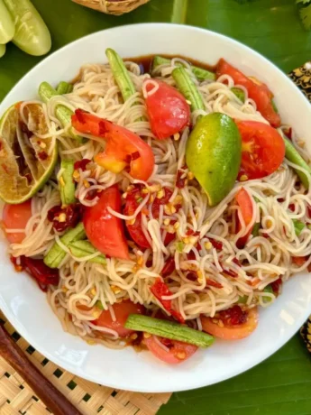Thum khao poon rice vermicelli salad served on a white plate with tomatoes, yard long beans, and a lime wedge, on a traditional Lao textile background.