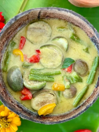 Vegetarian Thai green curry served in a coconut shell with green beans, eggplant, and bell peppers, surrounded by banana leaves and red and yellow flowers.