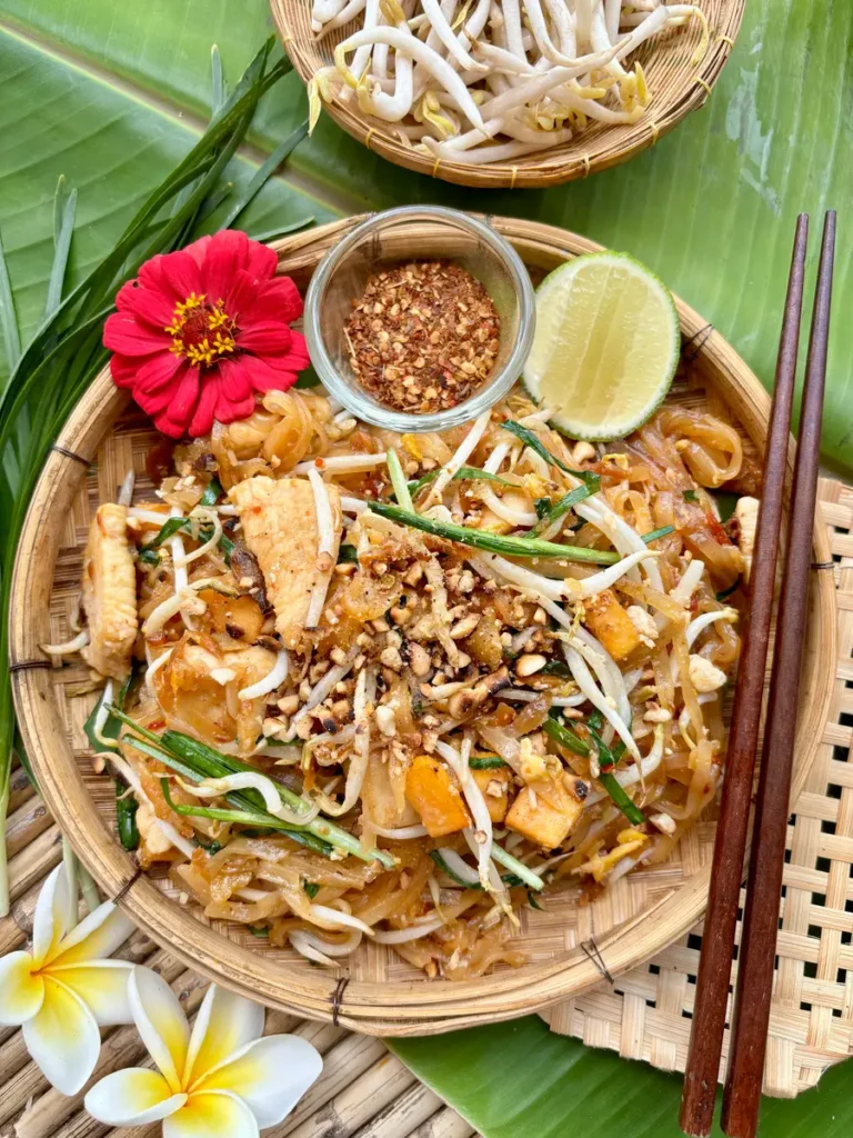 Authentic chicken pad Thai served in a traditional bamboo dish, accompanied by chili flakes and a lime wedge.