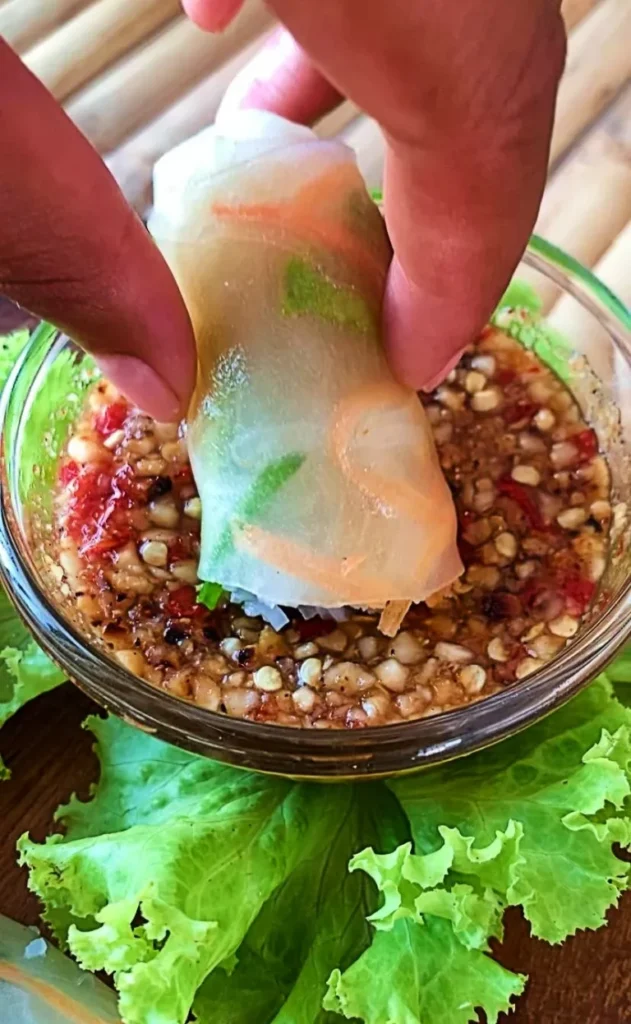 Hand dipping a Thai fresh spring roll into a bowl of spicy peanut sauce garnished with crushed peanuts and chili flakes.