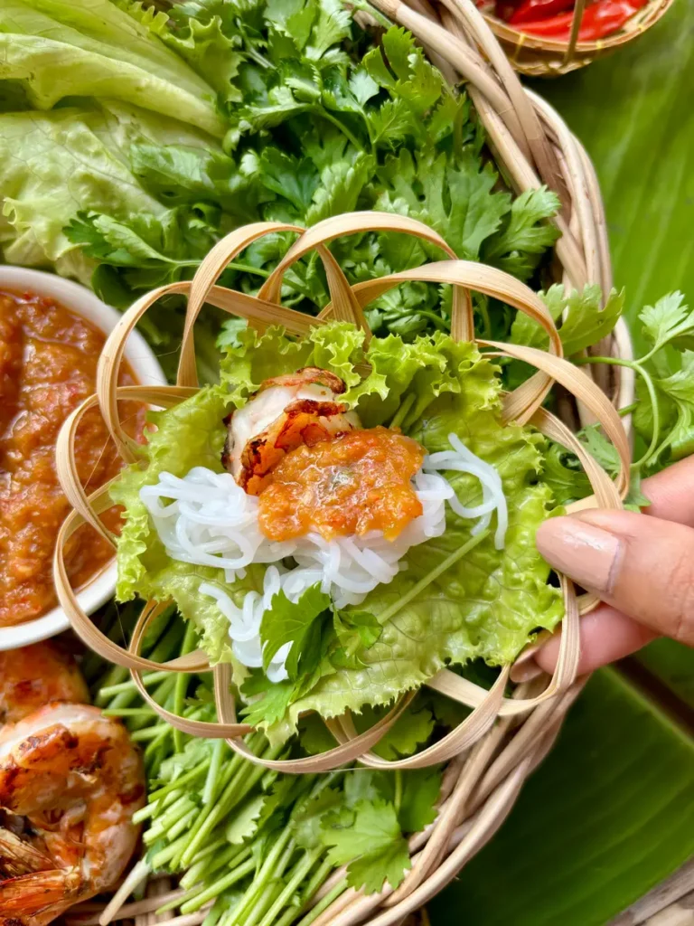 Hand holding bamboo basket with grilled Thai shrimps lettuce wrap over a plate with spicy peanut sauce and fresh vegetables.