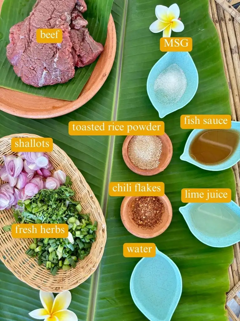 Ingredients for Thai ground beef larb displayed on banana leaves: raw beef, toasted rice powder, shallots, fresh herbs, chili flakes, lime juice, fish sauce, MSG, and water.