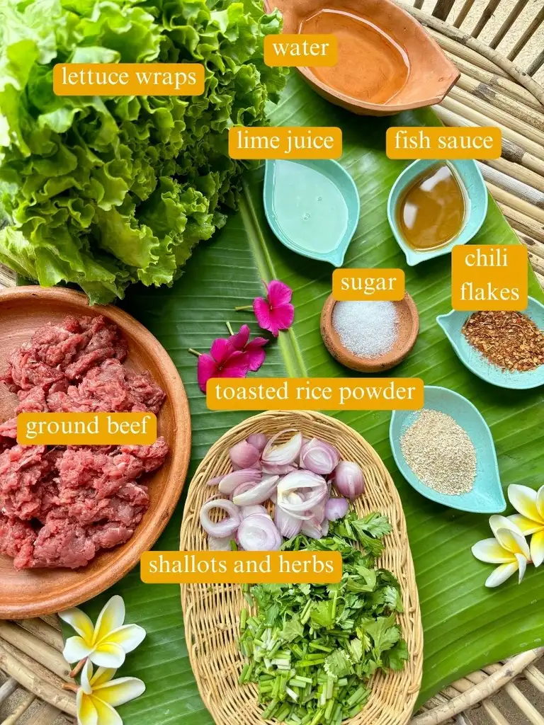 Top-view of ingredients for lettuce wraps: Lettuce, water, lime juice, fish sauce, sugar, chili flakes, toasted rice powder, shallots, fresh herbs, and ground beef.