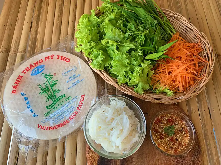Ingredients for Thai fresh spring rolls including Vietnamese rice paper, shredded carrots, fresh herbs, lettuce, rice noodles, and spicy dipping sauce.