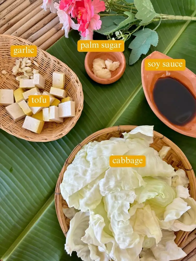 Ingredients for vegetarian cabbage stir-fry recipe: garlic, tofu, soy sauce, palm sugar, and cabbage, arranged on a banana leaf in clay cups and bamboo dishes.