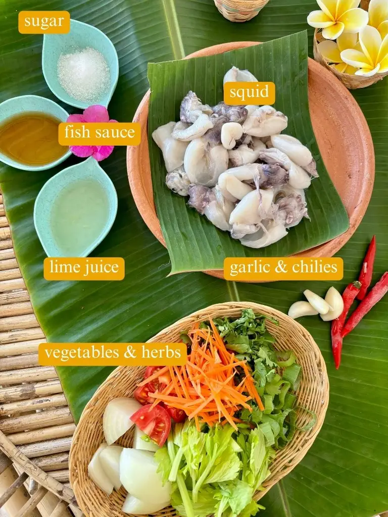 Ingredients for yum pla muk recipe laid out on a banana leaf: white sugar, fish sauce, squid, lime juice, garlic & chilies, vegetables & herbs. 
