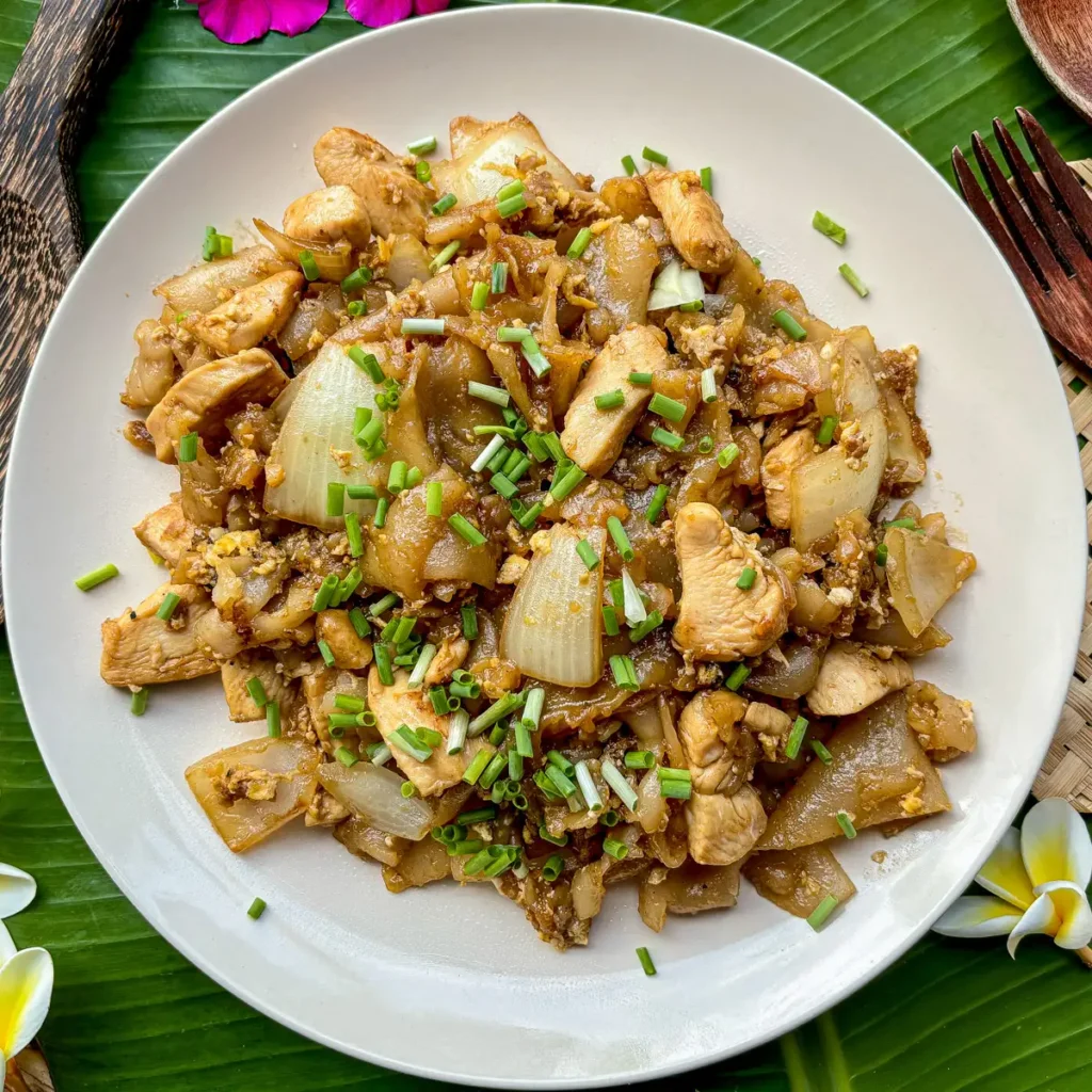 Overhead view of kuay teow kua gai stir-fry with tender chicken, wide noodles, and green onion on a white plate.