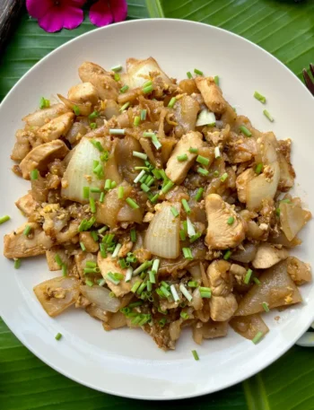 Kuay teow kua gai served in a white dish, showcasing pieces of roasted chicken and wide rice noodles garnished with green onions.