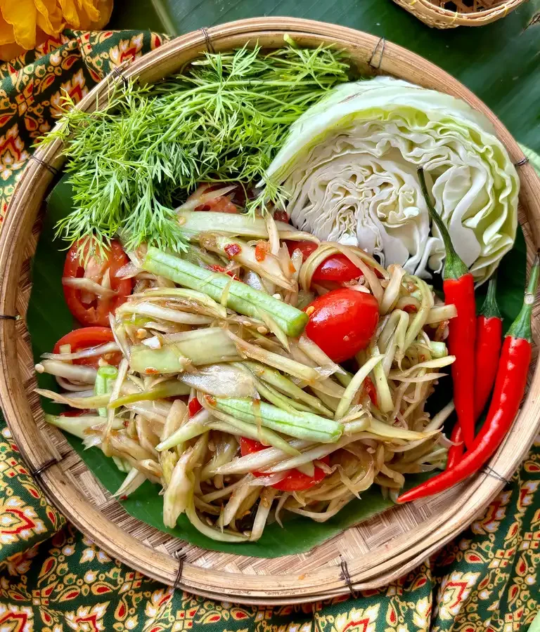 Traditional Lao papaya salad in a rustic serving basket, also known as thum mak hoong, with a side of fresh vegetables.