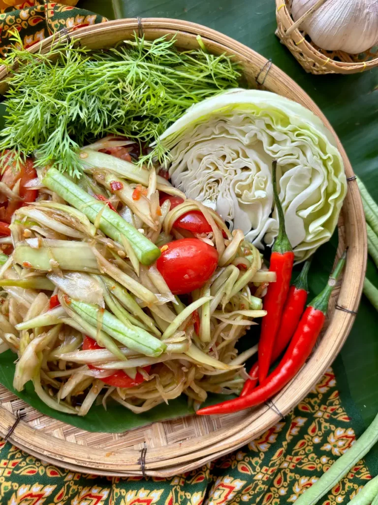 Freshly prepared Lao papaya salad in a woven basket, showcasing shredded green papaya, cherry tomatoes, long beans, and chili, garnished with lime and dill.