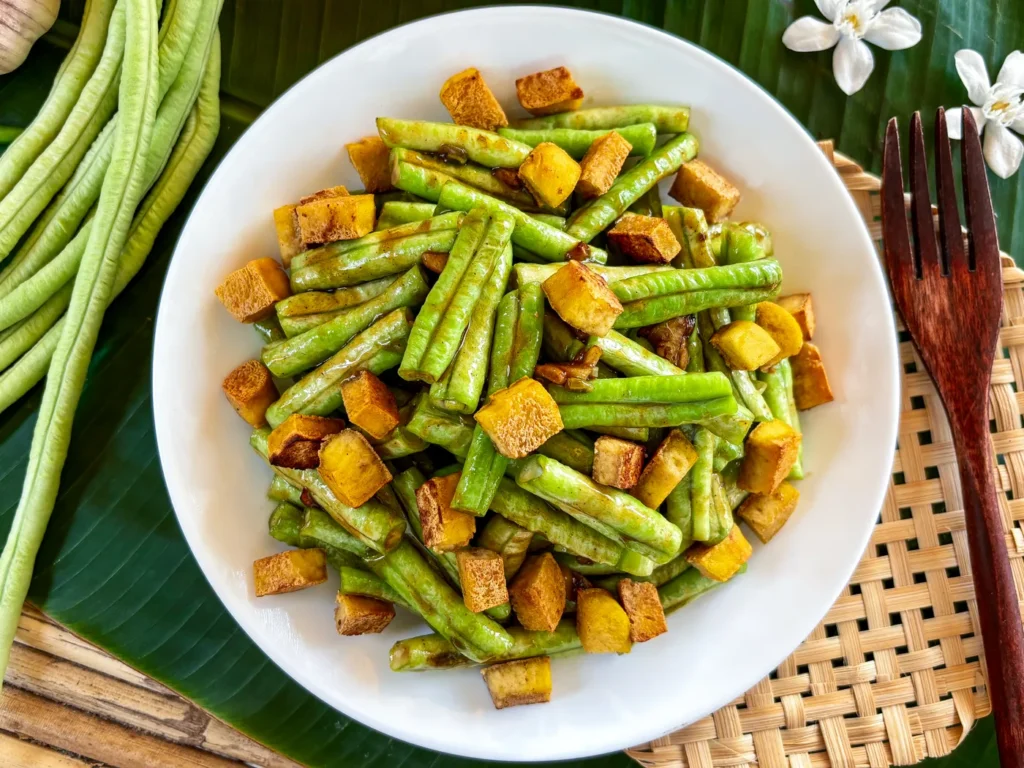 Long beans stir-fry with crispy tofu cubes served in a white bowl on a banana leaf background.