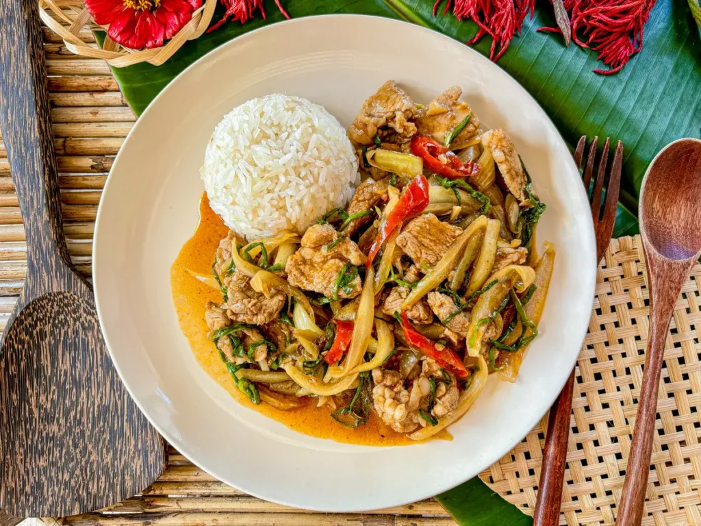 Thai moo pad prik pao, a spicy pork stir-fry, served with jasmine rice on a white plate, garnished with red chilies and green onions.
