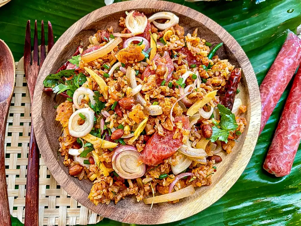 Nam khao tod Thai crispy rice salad with fresh herbs and sour sausage on a wooden plate over banana leaf.