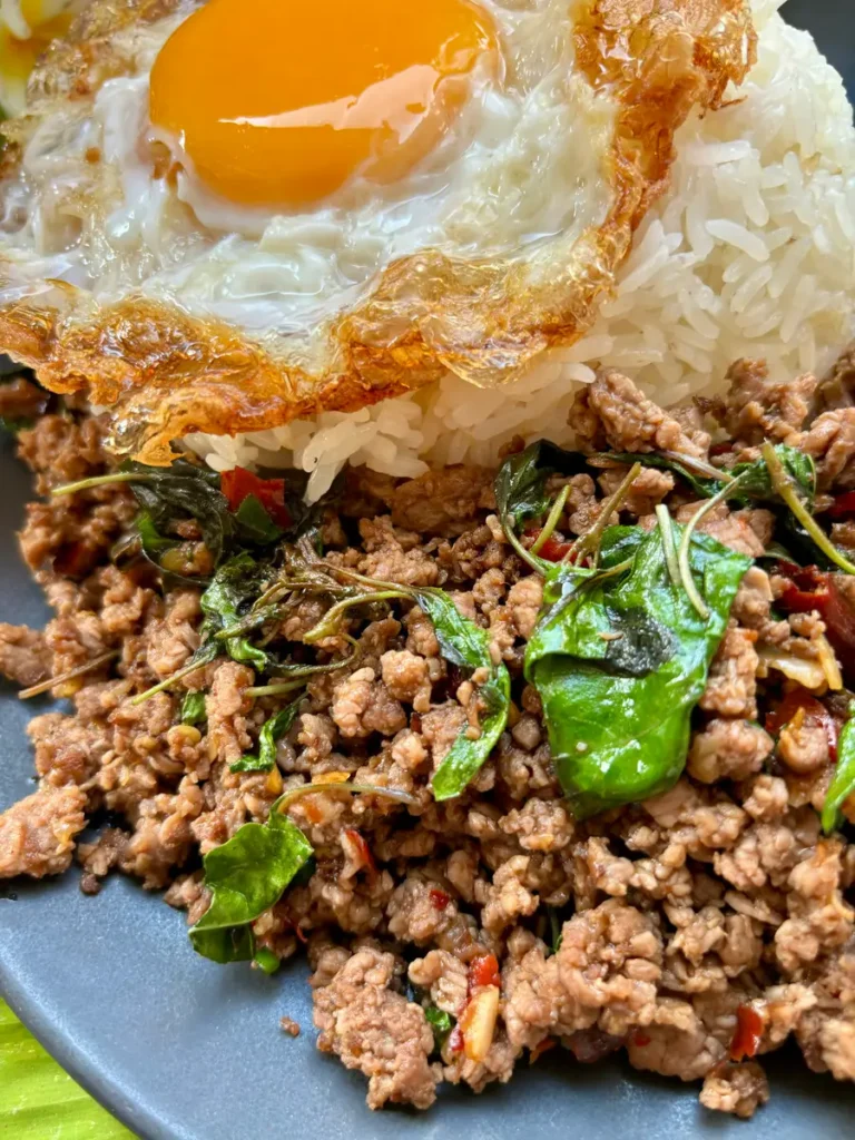 Suny-side-up egg over pad ga prow, Thai basil beef, with steamed rice on a plate.